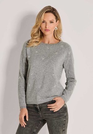 Frau mit blonden Haaren, die einen grauen Pullover mit kleinen Perlendetails und eine dunkelgraue Jeans trägt, posiert mit einer Hand in der Tasche vor einem schlichten Hintergrund.