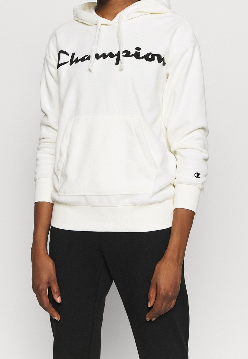Sweat à capuche crème avec une poche kangourou, logo « Champion » en écriture cursive noire. Comprend des cordons de serrage et des poignets et un ourlet côtelés. Associé à un pantalon noir.