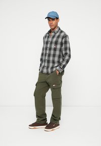 Juodas ir baltas languotas marškinėliai, alyvuogių žalumos kargo kelnės, mėlyna kepurė ir rudi sportbačiai su baltais akcentais; marškinėliai su logotipo etikete.