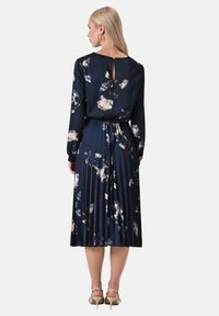 Marineblaues, floral bedrucktes Plisseekleid mit langen Ärmeln, Rückenausschnitt und weichem Stoff, der hellbeige und weiße Blumenmuster zeigt.
