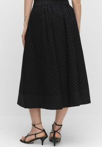Jupe midi noire avec un motif de points texturé, taille cintrée et coupe en A fluide. Associée à des sandales à talons avec lanières noires.