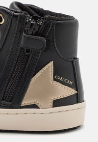 Musta nahkasaapas, jossa on sivuvetoketju ja vaaleanbeige tähtikoriste. Varusteena kultaiset yksityiskohdat ja vaaleasävyinen pohja. Tähti on painettu "GEOX"-logolla.