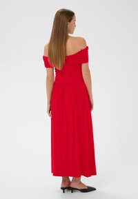 Robe maxi rouge à épaules dénudées avec des accents de tissu froncé, jupe fluide et corsage ajusté, accompagnée de mules à talons noirs.