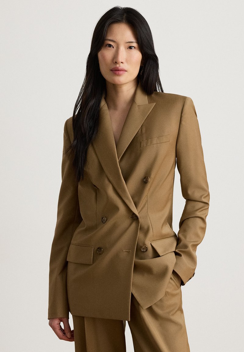 Γυναίκα με μακριά μαύρα μαλλιά που φοράει καφέ διπλόστενο σακάκι και αντίστοιχο παντελόνι, στεκόμενη με το ένα χέρι στην τσέπη μπροστά σε μονόχρωμο φόντο.