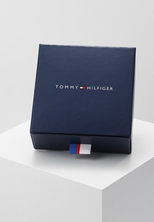 Boîte bleu marine Tommy Hilfiger avec logo et languette en tissu rouge, blanc et bleu de la marque sur un piédestal blanc.