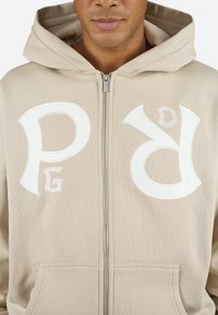 Beige zip-up hettegenser med store hvite broderte bokstaver "P" og "D" på brystet, med teksturert stoff og frontlomme.