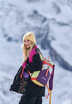 Kolorowy plecak z fioletowymi, zielonymi i pomarańczowymi akcentami; pikowany czarny płaszcz w kwiatowy wzór; żółta dzianinowa czapka. Zaśnieżone tło.