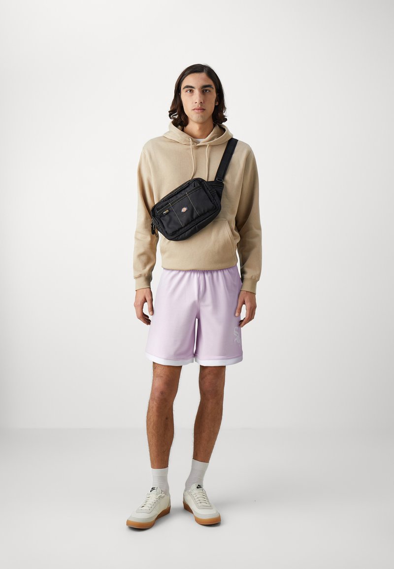 Sacoche noire porté en bandoulière avec plusieurs compartiments zippés, portée sur un hoodie beige et un short violet clair. Les baskets ont une semelle blanche et gomme.