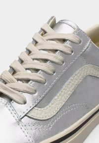 Ασημένιο και γκρι αθλητικό παπούτσι με λευκή υφή πλευρική ρίγα, κορδόνια εκρού και ορατή ραφή σε λείες και σουέτ επιφάνειες.