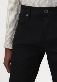 Svarta denimbyxor med klassisk passform, femficksdesign, knappstängning och kontrasterande sömmar mot en texturerad, ljusfärgad tröja.