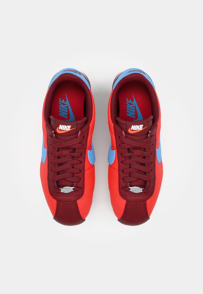 Zapatillas Nike con un vibrante upper rojo y acentos azules, acabado en tejido texturizado, cordones tradicionales y un detalle plateado en la punta.