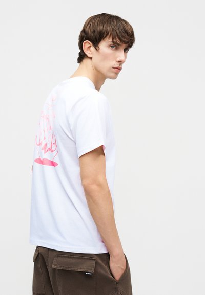 Νεαρός άνδρας με άσπρο Τ-shirt με ροζ αφηρημένο σχέδιο στην πλάτη, στέκεται με τα χέρια στις τσέπες, κοιτάζοντας πάνω από τον ώμο με φόντο μια απλή επιφάνεια.