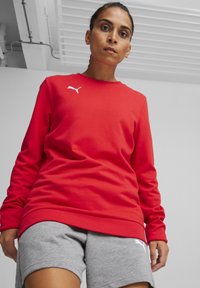 Felpa rossa a maniche lunghe con orlo e polsini a coste, con un logo bianco sul lato sinistro del petto. Abbinata a pantaloni corti grigi.