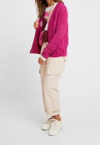Femme portant un cardigan rose vif par-dessus un pull multicolore, un pantalon cargo beige et des baskets à plateforme couleur crème.