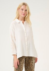 White button-up shirt in textured fabric, featuring long sleeves and light embroidery on the shoulders. Paired with leopard-print pants.