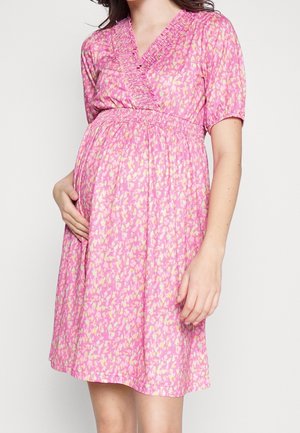 Schwangere Frau trägt ein knieumspielendes Kleid mit rosa Muster und hält sanft ihren Bauch mit einer Hand.