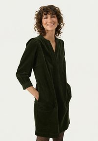 Dunkelgrünes Cordkleid mit V-Ausschnitt, Dreiviertelärmel, seitlichen Taschen und strukturierten vertikalen Streifen.