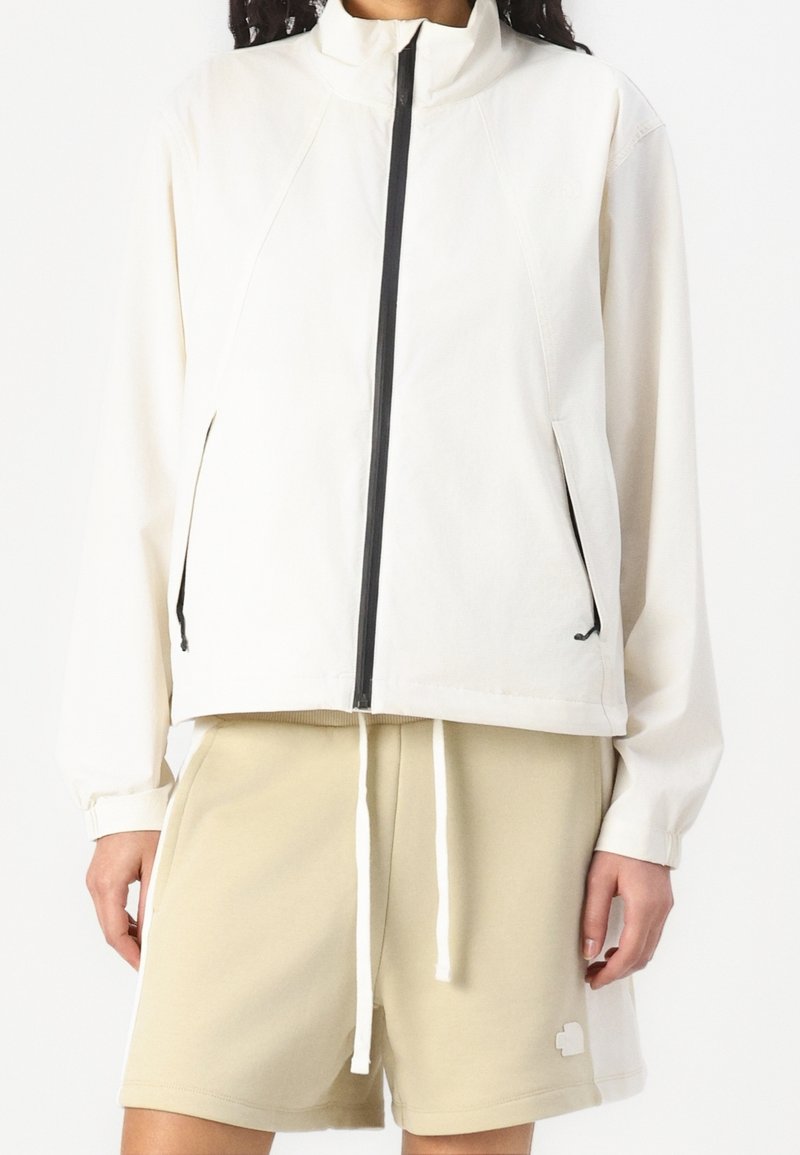 Personne portant une veste crème à fermeture éclair noire et un short beige à cordon, sur un fond blanc uni.