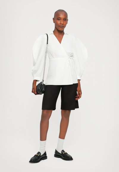 Witte wikkelblouse met puffmouwen, gecombineerd met zwarte bermuda-shorts. Accessoires omvatten een kleine zwarte tas en zwarte loafers met gouden accenten.