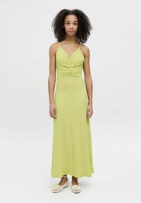 Robe maxi en tissu vert clair texturé, dotée de fines bretelles et d'un décolleté en V. La robe se resserre au niveau de la poitrine, créant une silhouette épurée.