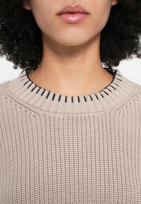 Gaiši bēša adīta džempera ar ribotu rakstu. Apkakle izcelta ar tumši zilu šuvi, apkārt apkaklei ir smalkas detaļas.