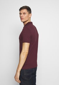 Bordeaux polo-shirt met korte mouwen, geribbelde kraag en rechte zoom. Gebruikt in combinatie met donkere jeans, weergegeven vanuit een zijaanzicht.