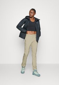 Schwarze Pufferjacke mit Kapuze, kombiniert mit einem schwarzen Cropped-Top und beigen, abnehmbaren Hosen. Die Schuhe haben blaue und gelbe Akzente.