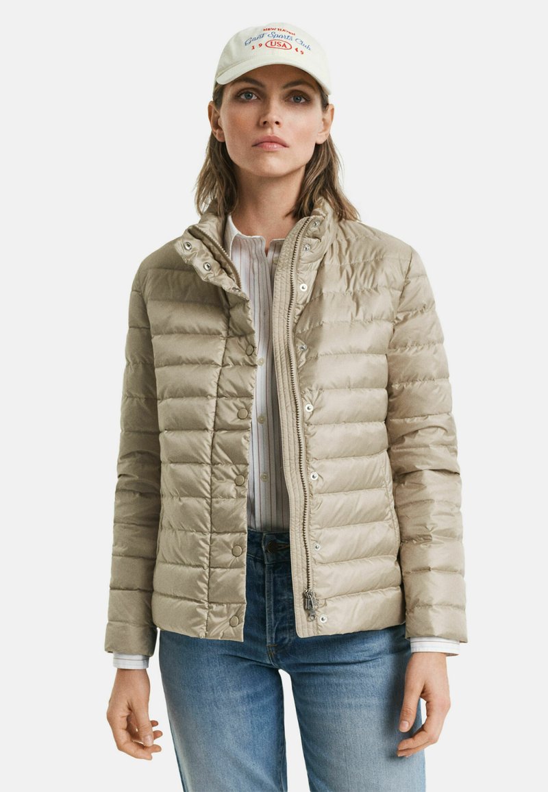 Veste doudoune beige avec un design matelassé, boutons-pression et fermeture éclair sur le devant. Portée sur une chemise blanche à rayures, associée à un jean bleu.