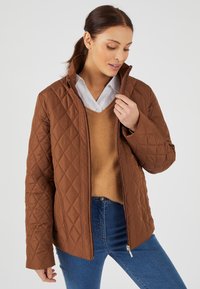 Giubbotto marrone trapuntato con un colletto alto, tasche laterali con zip e una texture liscia. Indossato sopra un maglione marrone chiaro e jeans blu.