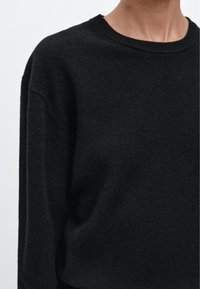 Schwarzer Strickpullover mit rundem Ausschnitt, fallenden Schultern und weicher Textur. Der Stoff wirkt leicht und gemütlich.