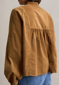 Veste en velours côtelé marron avec une finition texturée, détail de dos froncé et manches longues volumineuses. Col légèrement structuré.