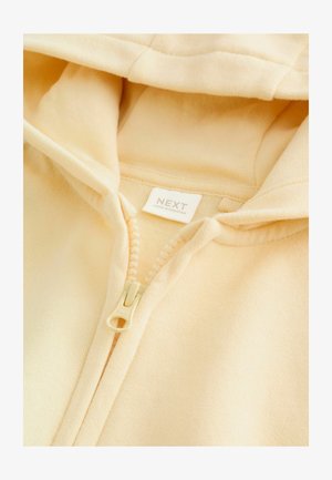 Svetložltá mikina na zips má mäkkú látku, klokanie vrecko a bielu etiketu s nápisom "NEXT." Zips je krémovej farby.