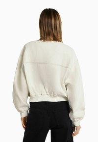 Zkrácená béžová mikina vyrobená z měkké tkaniny s spadenými rameny, žebrovanými manžetami a pasem, s viditelným švem na zadní straně.