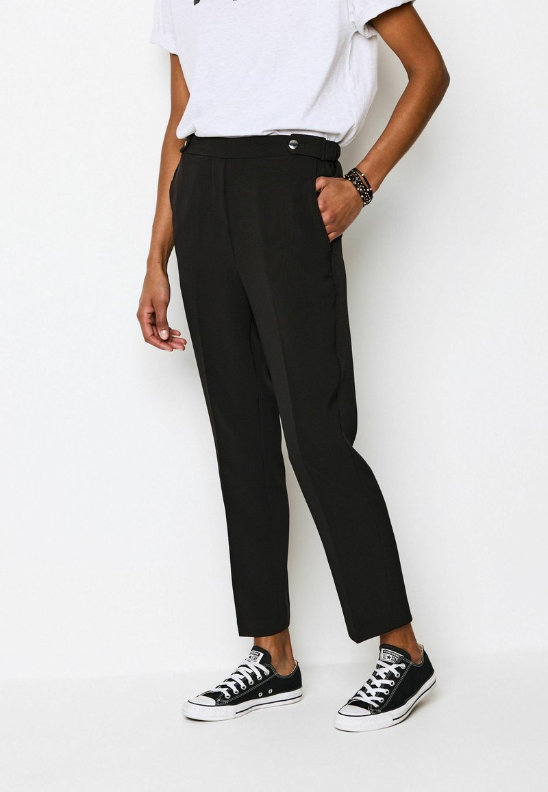 Schwarze, taillierte Hose mit hoher Taille, glatter Oberfläche, Seitentaschen und silbernen Details, kombiniert mit schwarzen Canvas-Sneakers.