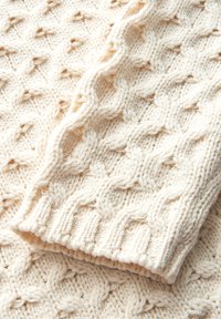 Tissu tricoté de couleur crème avec un motif de câble texturé. Le tissu présente un ourlet côtelé et une texture douce et moelleuse.