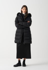 Piumino nero con cappuccio foderato in pelliccia, design a scatola, sezioni trapuntate, chiusura con zip, indossato sopra una gonna nera e mocassini.