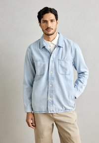 Light blue denim jacket with two front pockets, button closure, and a relaxed fit. Features a collar and seams with minimal stitching details.