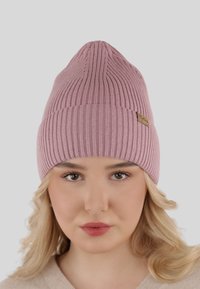 Pink knit beanie with ribbed texture, pointed top, and a small wooden logo tag on the side. Soft material and fitted design.