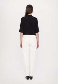 Geschnittenes schwarzes Shirt mit einem strukturierten Muster und weißen hochgeschnittenen Hosen. Schwarze Plateauschuhe vervollständigen das Outfit. Von hinten betrachtet.
