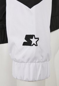Chaqueta en blanco y negro que presenta una manga negra con un puño elástico y un logo bordado prominente en blanco y negro.