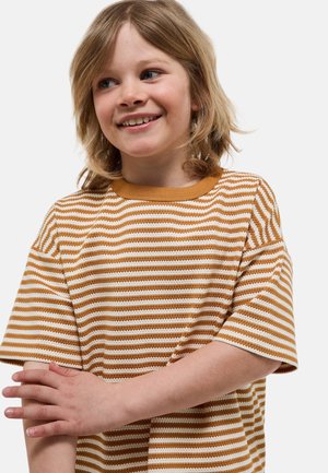 Enfant souriant aux cheveux blonds mi-longs portant un t-shirt à manches courtes rayé moutarde et blanc, les bras croisés à la taille.