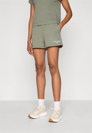 Grønne bomuldsshorts med en afslappet pasform, udstyret med elastisk talje og hvid logo detalje på kanten. Kombineret med lette sneakers og sokker.