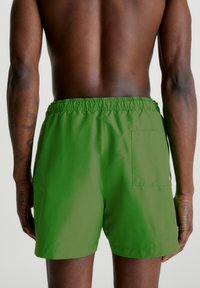 Grønne badetights laget av lett stoff, med elastisk midje, sidelommer og en baklomme med trykknapp.