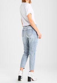 Bílé tričko s krátkým rukávem, světle modré vybledlé džíny ve stylu mom a bílé kotníkové boty na podpatku se zlatými cvočky, pohled zezadu.