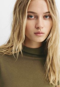 Olivovo zelený rolák, detail tváre s svetlými blond vlasmi, čistou pleťou a prirodzeným make-upom, oči sa pozerajú priamo na diváka.