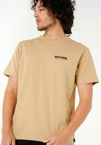 Tan katoen t-shirt met korte mouwen; heeft een zwart en rood logo op de linkerborst. Zachte textuur, reguliere pasvorm en ronde hals.