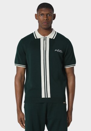 Man draagt een donkergroene polo met korte mouwen, verticale witte strepen en een witte kraag, kijkt recht vooruit tegen een effen lichte achtergrond.