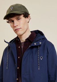 Marineblauer Windbreaker mit Kapuze, ausgestattet mit einem Logo auf der Vorderseite und einem gemusterten Kordelzug. Kombiniert mit einer olivgrünen Cordmütze. Das Modell hat einen neutralen Hintergrund.