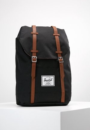 Sac à dos Herschel noir avec des sangles en cuir marron et des boucles argentées, debout sur une surface blanche contre un fond uni.
