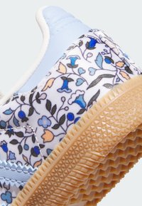 Blaue und weiße Sneaker mit floralen Mustern, strukturiertem Stoff und einer Gummisohle mit gemustertem Profil. Akzente in Blau, Gelb und Grün.